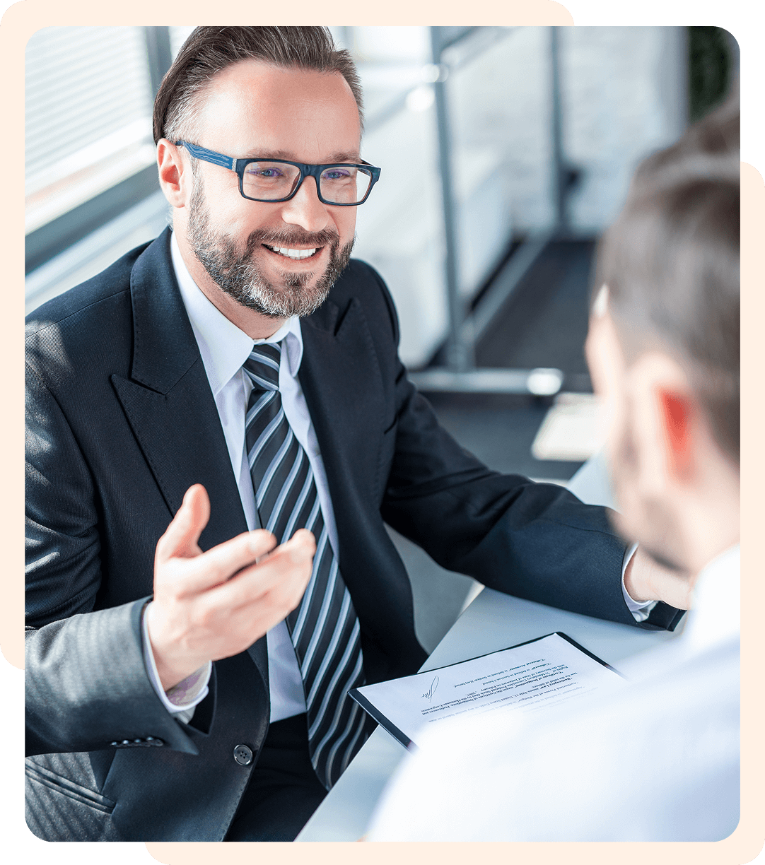 2Group 1321315022 A man in a suit and glasses is sitting at a desk, gesturing with his hand, conversing with another person. A document is on the table.